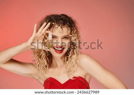overjoyed woman with curly hair and red lips holding makeup foundation on pink and yellow backdrop