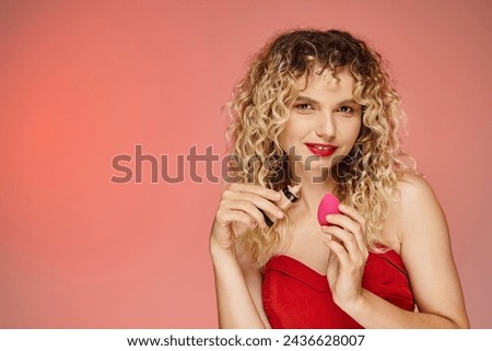 attractive curly woman holding cosmetic sponge and makeup foundation on pink and yellow backdrop