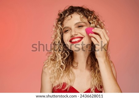 curly cheerful woman with red lips using face sponge on pink and yellow backdrop, elegant beauty