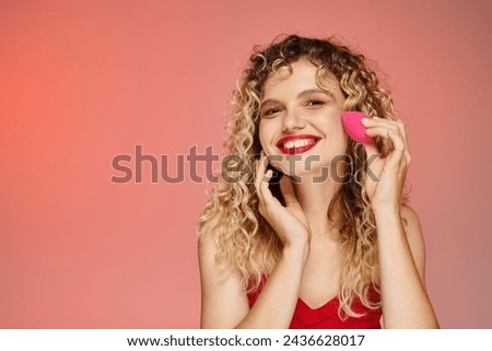 curly joyful model with red lips using face sponge on pink and yellow backdrop, elegant beauty