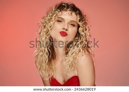 flirty curly woman with bold makeup pouting lips while sending air kiss at camera on pastel backdrop