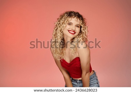 alluring woman with bold makeup and wavy hair posing in red top looking at camera on pastel backdrop