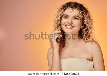 cheerful woman with curly hair and perfect skin using jade roller on pink and yellow backdrop