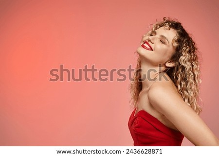 delighted woman with wavy hair posing in red top with closed eyes on pink and yellow backdrop