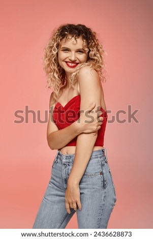 jolly woman with red lips and curly hair posing in red top and smiling at camera on pastel backdrop