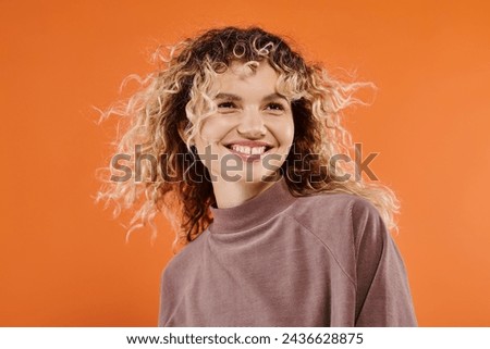 carefree woman with curly hair in mocha color turtleneck looking at camera on bright orange backdrop