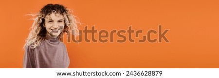 joyful woman with wavy hair in mocha color turtleneck looking at camera on orange backdrop, banner