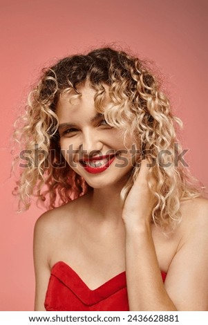 portrait of excited woman with curly hair and red top smiling at camera on pastel pink backdrop