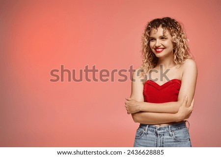 cheerful smiling woman with wavy hair in red top looking away on pink and yellow backdrop