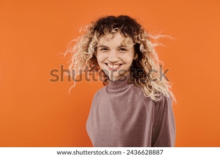 cheerful woman with wavy hair in mocha color turtleneck looking at camera on radiant orange backdrop