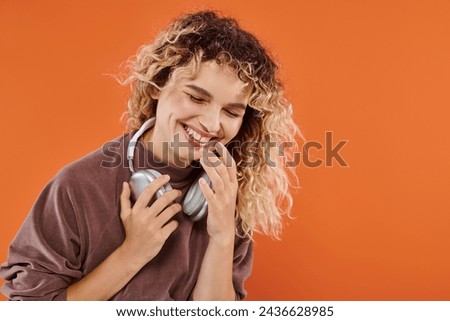 excited curly woman in brown turtleneck with wireless headphones laughing on orange backdrop