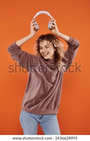 overjoyed curly woman in stylish turtleneck dancing with wireless headphones on orange backdrop