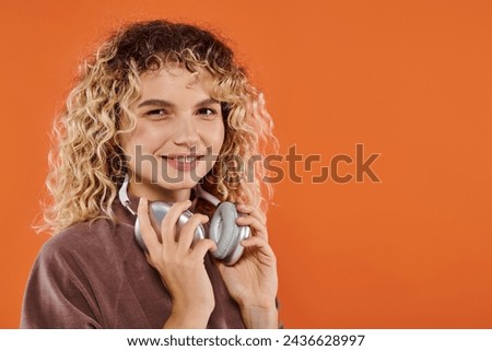 positive curly woman in trendy turtleneck with wireless headphones looking at camera on orange