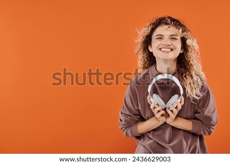 excited curly woman in brown turtleneck posing with wireless headphones on orange backdrop