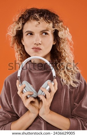 thoughtful curly woman in brown turtleneck holding wireless headphones and looking away on orange