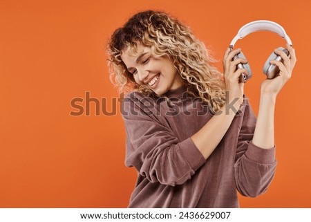 carefree and stylish curly woman in mocha color turtleneck holding wireless headphones on orange