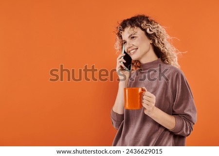 cheerful curly woman with cup of morning coffee talking on smartphone on bright orange backdrop