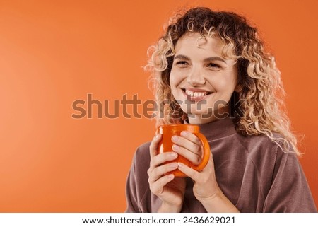 cheerful woman with curly hair and cup of morning coffee looking away on orange, daily routine
