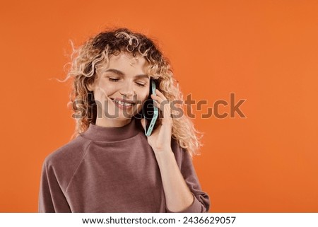 smiling woman with curly hair in brown turtleneck talking on smartphone on radiant orange backdrop