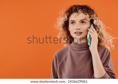 joyful woman with wavy hair in brown turtleneck talking on mobile phone on radiant orange backdrop