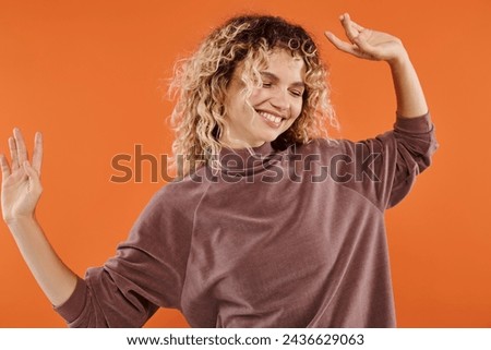 jolly woman with wavy hair in stylish mocha color turtleneck posing on vibrant orange backdrop
