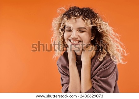 trendy woman with wavy hair in mocha color turtleneck smiling with closed eyes on orange backdrop
