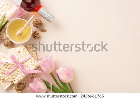 Top view picture of a Passover seder arrangement with matzah in a ribbon, a red wine bottle, walnuts, bowl of honey with a dipper spoon, and fresh pink tulips, on a pastel beige surface