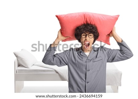 Angry young man in pajamas holding a pillow over head and screaming in front of his bed isolated on white background