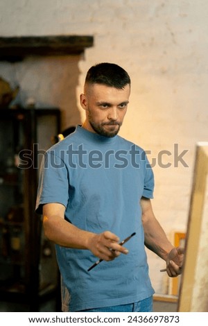 in an art workshop an artist in a blue T-shirt measures with a brush on canvas and thinks