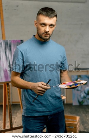 in an art studio an artist stands directly at the camera with a palette and smiles