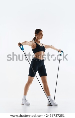 Fit young woman in black sportswear training, using resistance bands for arm exercise isolated over white studio background. Concept of sport, health and body care, fitness app, exercises templates