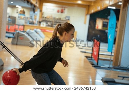 Side view of a young long-haired blonde girl dressed in black throwing the bowling ball onto the lane
