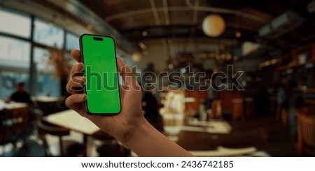 Person holding a smartphone with a green screen in a blurred cafe setting, perfect for showcasing apps and mobile technology