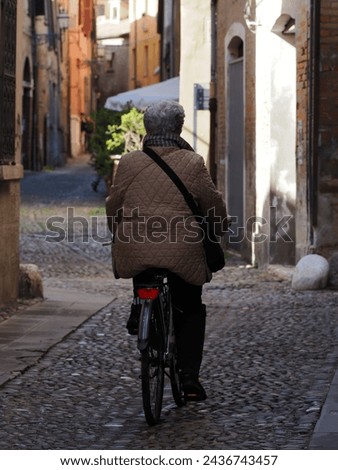 Ferrara, Italy. Elderly woman on a bicycle in a narrow alley in the historic center. Royalty-Free Stock Photo #2436743457