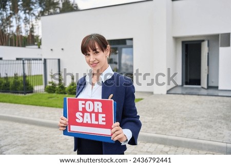 Woman realtor stands near a country house with a for sale sign