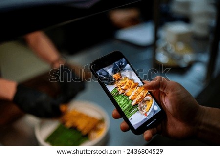 A person captures a vibrant photo of delicious grilled food on a smartphone. The fresh meal, displayed on green leaves, is beautifully showcased. The indoor setting suggests a kitchen or dining area