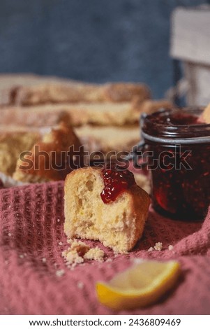 close-up image of a cupcake with a glass jar of raspberry or strawberry jam. it is on a wooden table with slices of bread and a pink cloth.