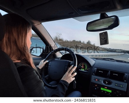Young blonde long-haired girl driving the grand tourer car at sunset on a long trip.Interior view, good driver, attentive and with her hands on the wheel. Dusk with reddish sky Royalty-Free Stock Photo #2436811831