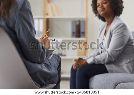Female psychologist or therapist with clipboard and pencil in hands sitting on chair and talking to young African American woman patient on sofa. Therapy, psychotherapy concept Royalty-Free Stock Photo #2436813051