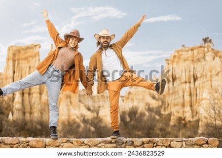 A man and woman are posing for a picture in front of a mountain. The man is wearing a cowboy hat and the woman is wearing a hat and jeans. They are both smiling and holding hands
