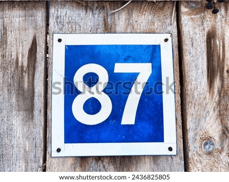 Number 87 on a Blue Sign Against Wooden Background. A bold white number 87 centered on weathered blue square sign