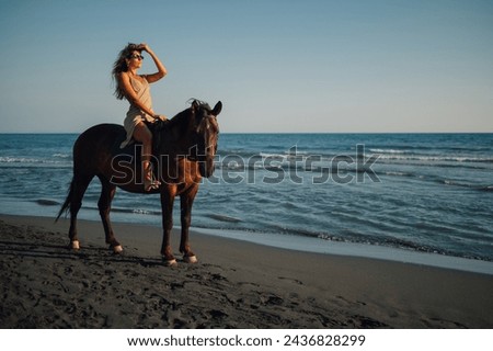 Portrait of a gorgeous woman horseback riding on a horse while looking at beautiful and scenic sunset on a beach near water. Dreamlike picture of a young lady riding a horse on a beach near ocean.