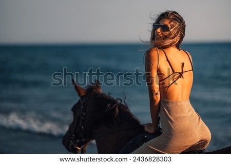 Portrait of a young attractive tattooed woman riding a horse at sunset on a beach near the sea or ocean while looking over her shoulder and smiling at the camera. A lady is horseback riding at beach.