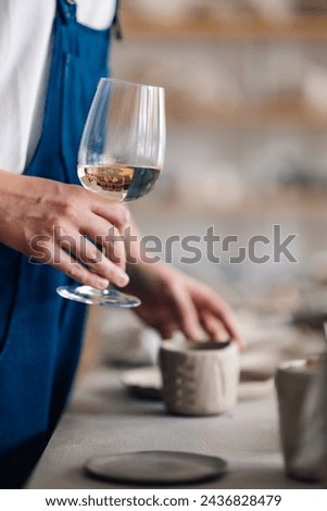 Cropped picture of a pottery designer's hand holding a wineglass at ceramics studio. Pottery and wine concept. Close up of a craftswoman having a glass of wine after pottery class at creative workshop