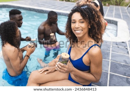 Biracial woman in blue swimwear enjoys a drink by the pool; diverse friends chat in the background. It is a sunny day, perfect for outdoor relaxation and socializing.
