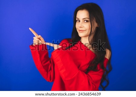 Look, advertise here! Dark haired pretty woman with appealing smile gives positive attitude towards good deal, expresses her recommendation while pointing on left side isolated on blue background. Royalty-Free Stock Photo #2436854639