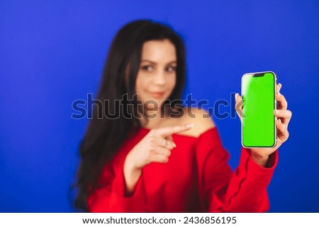 Excited young woman in red top hold and show blank phone green screen with copy space for app, isolated on blue background. Happy woman with wow emotions, recommend app or some product. Focus on phone