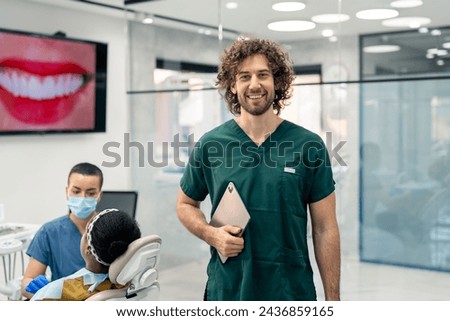 Handsome dentist in green uniform holding digital tablet in modern clinic with patient in dentist's chair and nurse in background. Healthy smile presentation on tv screen also visible in background.