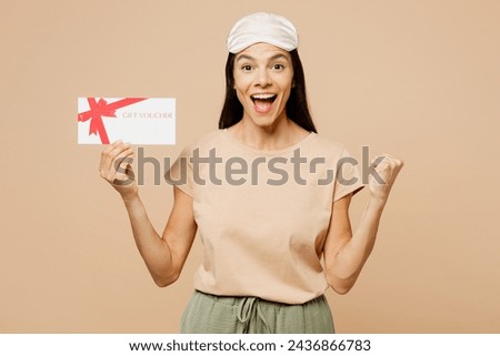 Young Latin woman wears pyjamas jam sleep eye mask rest relax at home hold gift coupon voucher card for store do winner gesture isolated on plain beige background studio. Good mood night nap concept