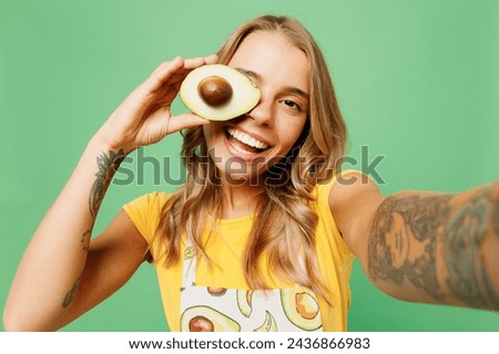 Close up young housewife housekeeper chef cook baker woman wear apron yellow t-shirt do selfie shot on mobile cell phone cover eye with avocado isolated on plain green background. Cooking food concept